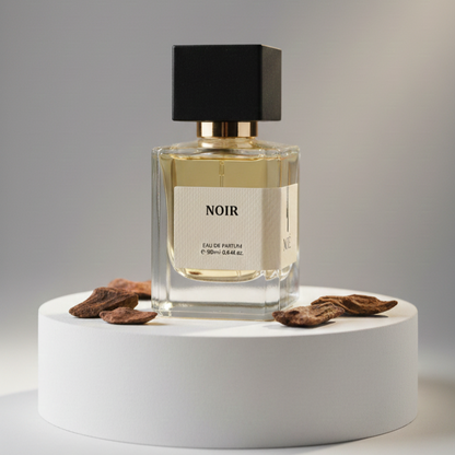 Square glass perfume bottle labeled Noir with black cap on white pedestal surrounded by dried brown petals