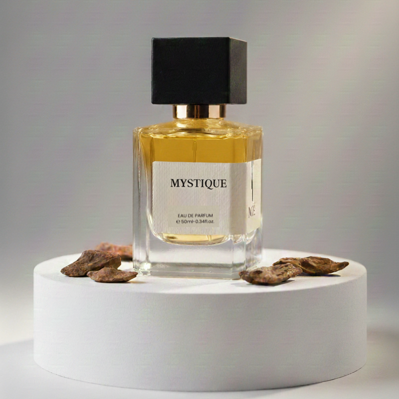 Mystique eau de parfum bottle with black cap on white round pedestal surrounded by dried brown flower petals