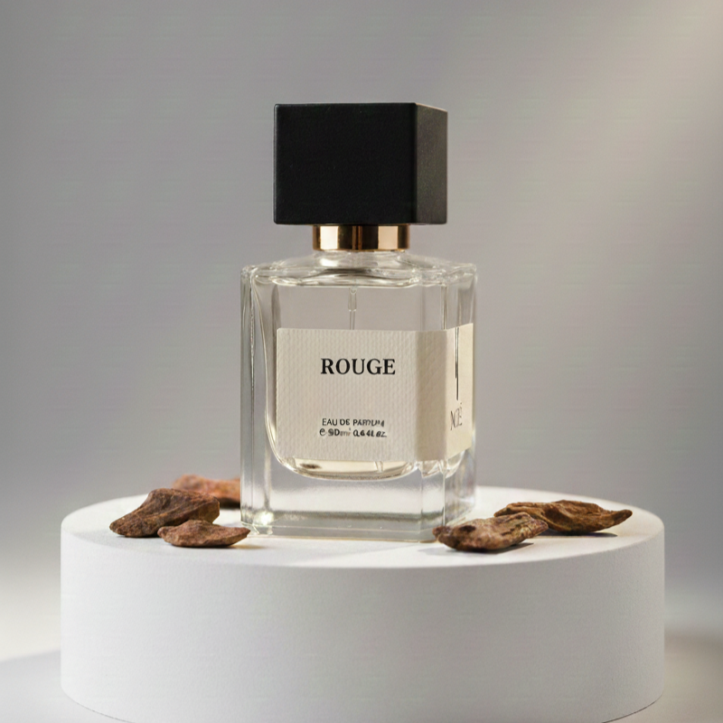 Clear glass perfume bottle labeled Rouge Eau de Parfum with black cap on white pedestal surrounded by dried wood pieces